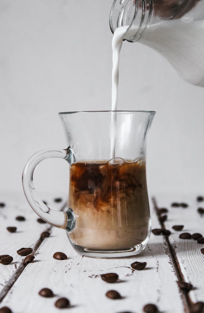 Glass mug of coffee with milk being poured, surrounded by scattered coffee beans on a wooden table.