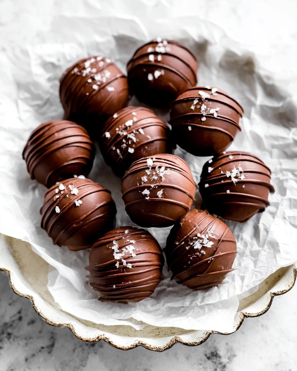 A group of round chocolate truffles sits on crumpled white parchment paper that covers a white scalloped metal tray. Each truffle is smooth dark brown with thin darker chocolate drizzle lines on top and small white salt flakes sprinkled over them, creating a textured contrast. The truffles are placed close together in a loose pile, showing their shiny, glossy surface under soft light. The whole scene is set on a white marbled texture background. photo taken with an iphone --ar 4:5 --v 7