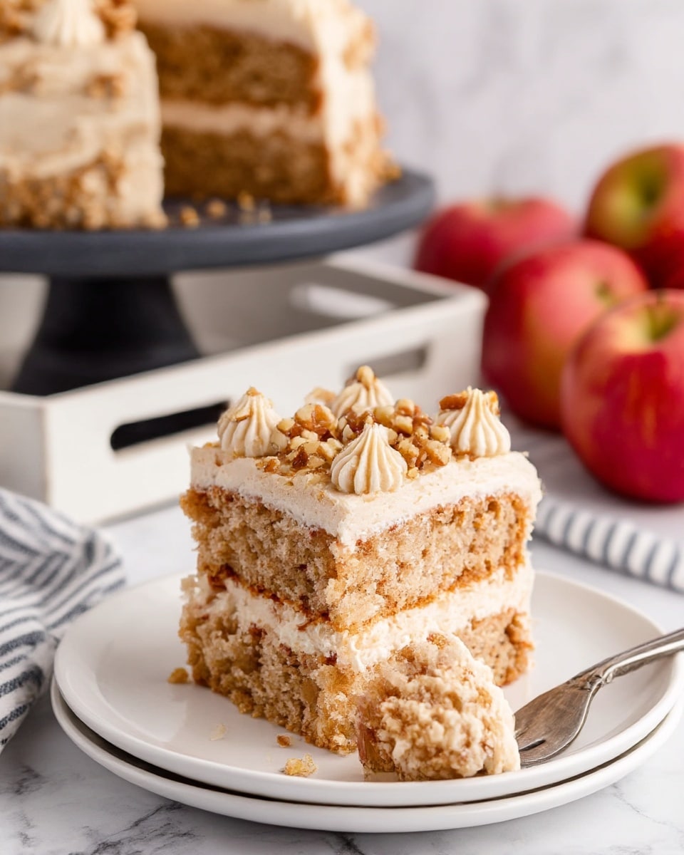A close-up of a two-layer square cake piece on a white plate, each light brown layer showing a crumbly, moist texture with small bits inside. Between the layers and on top, there is a thick layer of smooth light tan frosting, with some small dollops on the side. The top layer of frosting is sprinkled with chopped walnuts, adding a rough texture. A fork on the right side of the plate holds a small bite of the cake. The background includes a white marbled surface, a white tray with a striped cloth, three whole red apples, and a dark cake stand holding the rest of the cake, slightly blurred. photo taken with an iphone --ar 4:5 --v 7