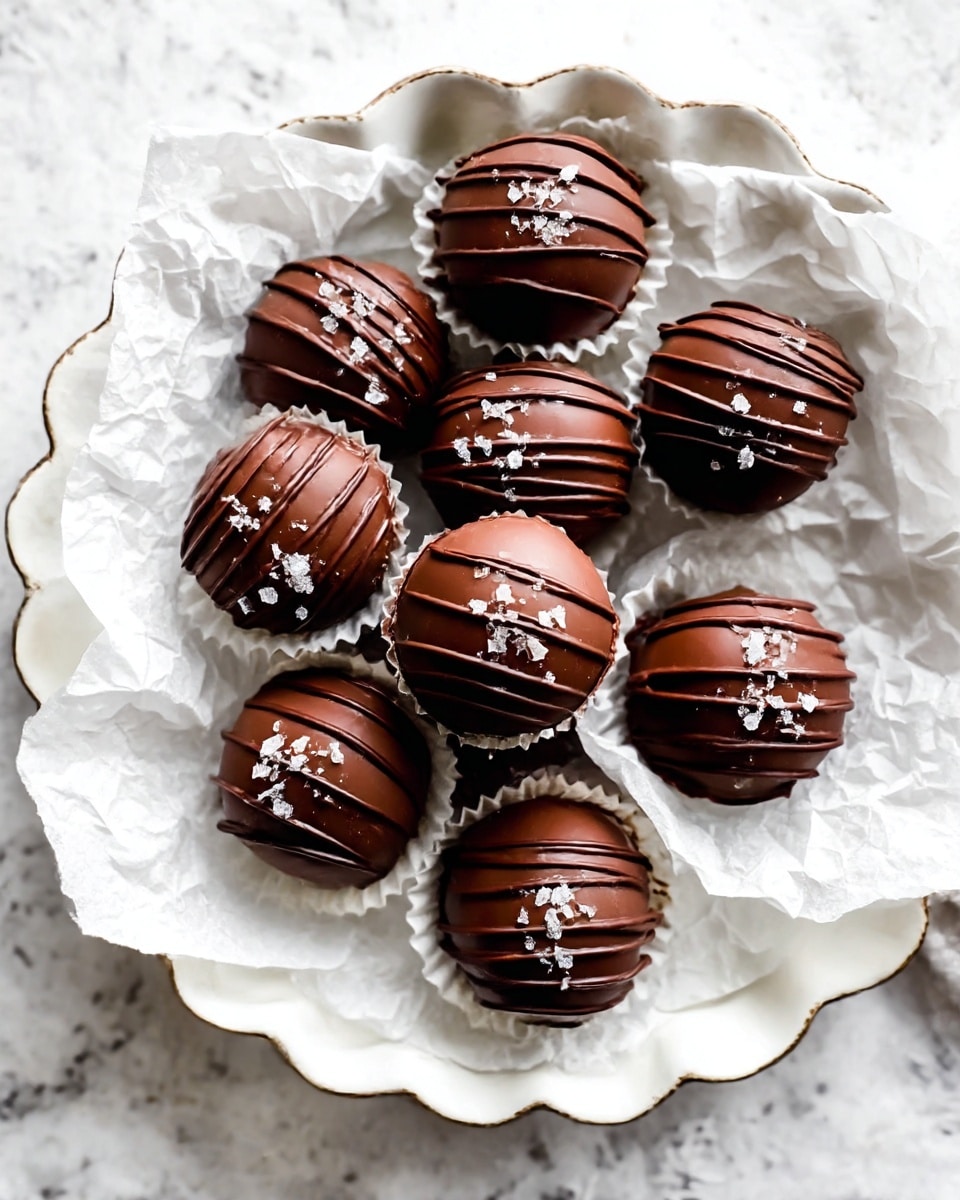 A group of small round chocolate truffles with a smooth dark brown shell are placed on crumpled white parchment paper inside a white scalloped-edge tray. Each truffle is decorated with thin dark chocolate drizzle lines and sprinkled with small white flakes of sea salt on top. The tray rests on a white marbled textured surface. photo taken with an iphone --ar 4:5 --v 7