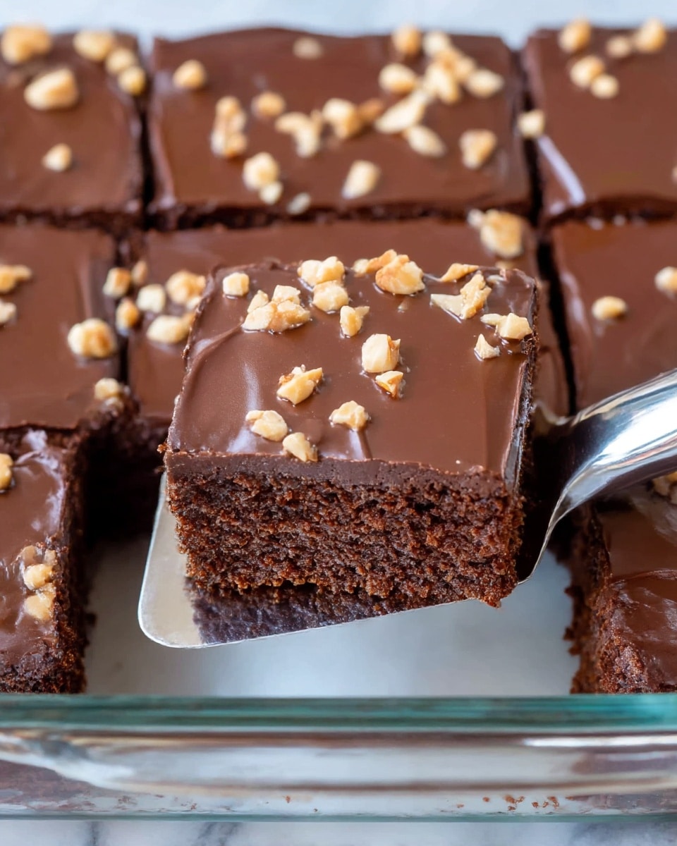 A close-up of a chocolate cake cut into square pieces with one piece being lifted by a metal spatula. The cake has two visible layers: a thick, moist dark brown base and a smooth, glossy dark chocolate frosting on top. The frosting is sprinkled with small, light tan chopped nuts, evenly scattered across the surface. The cake sits in a clear glass dish on a white marbled texture background. Photo taken with an iphone --ar 4:5 --v 7