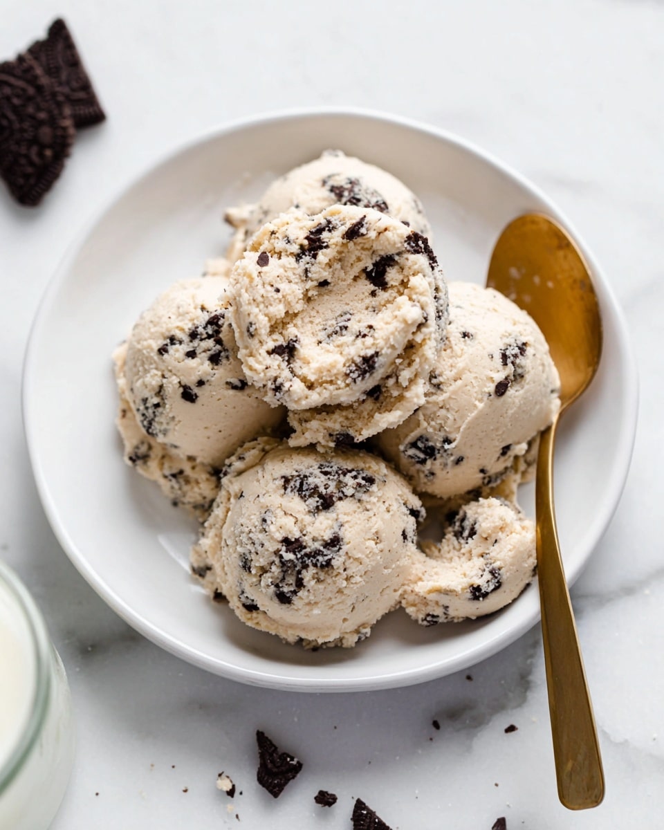 A white plate holds four scoops of light beige cream cookie dough mixed with dark cookie chunks, giving a rough, textured look. The scoops are piled closely together, with a golden spoon scooping some dough from the side. Around the plate on a white marbled background, broken pieces of the dark cookies with white cream filling are scattered, adding contrast. In the foreground, a blurry glass bottle filled with white liquid is partly visible, and in the back left, a stack of white plates is slightly out of focus. Photo taken with an iphone --ar 4:5 --v 7