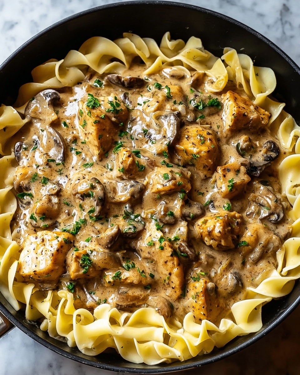 A black skillet holds a creamy chicken dish over egg noodles. The bottom layer consists of wide, pale yellow egg noodles with soft, ruffled edges. On top, a rich light brown sauce with visible specks of black pepper and herbs covers chunky pieces of golden-brown seared chicken and sliced dark brown mushrooms. The dish is sprinkled with finely chopped green parsley, adding a fresh contrast. The skillet is set on a white marbled surface. photo taken with an iphone --ar 4:5 --v 7