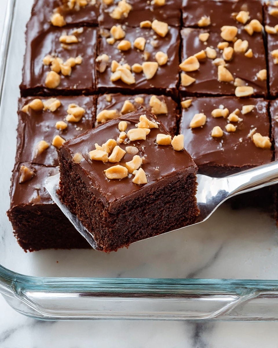 A clear glass baking dish holds a rich chocolate cake cut into squares, with one piece slightly lifted by a silver spatula from the right side of the dish. The cake has two layers: a moist, textured dark brown base and a smooth, glossy dark chocolate frosting on top. Scattered over the frosting are small pieces of light tan chopped peanuts, adding a crunchy contrast. The white marbled surface beneath the dish creates a clean and bright background. photo taken with an iphone --ar 4:5 --v 7