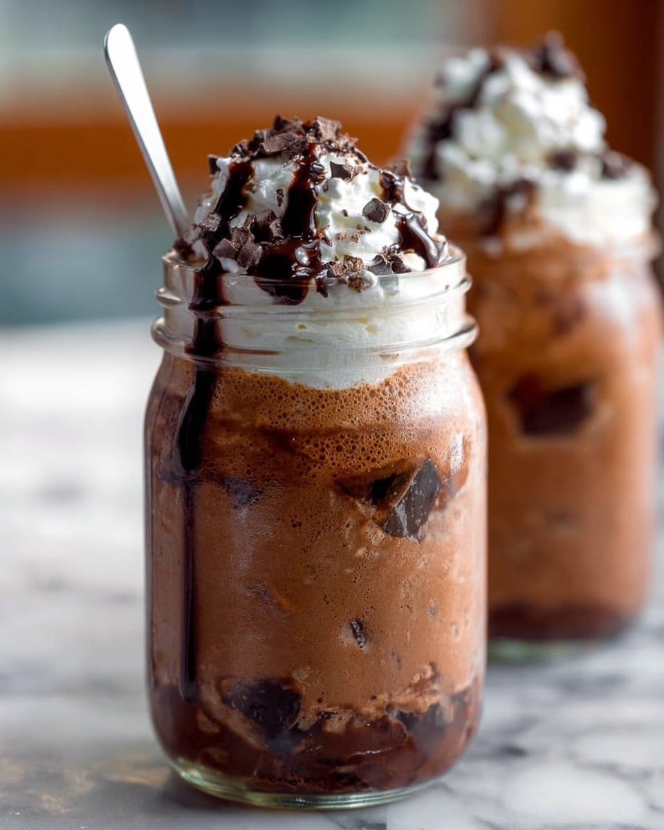 The image shows a cold dessert drink served in a clear glass jar, featuring three main layers: the bottom layer is a thick, brown chocolate mixture with smooth texture and some darker chocolate chunks, the middle layer contains more dense dark chocolate pieces embedded in the mix, while a creamy white whipped cream with a light fluffy texture tops the jar, drizzled with dark chocolate syrup that runs slightly down the sides of the jar. A white spoon is placed inside the jar near the whipped cream, and in the blurred background, there is another similar jar. The jars are on a white marbled surface. Photo taken with an iphone --ar 4:5 --v 7