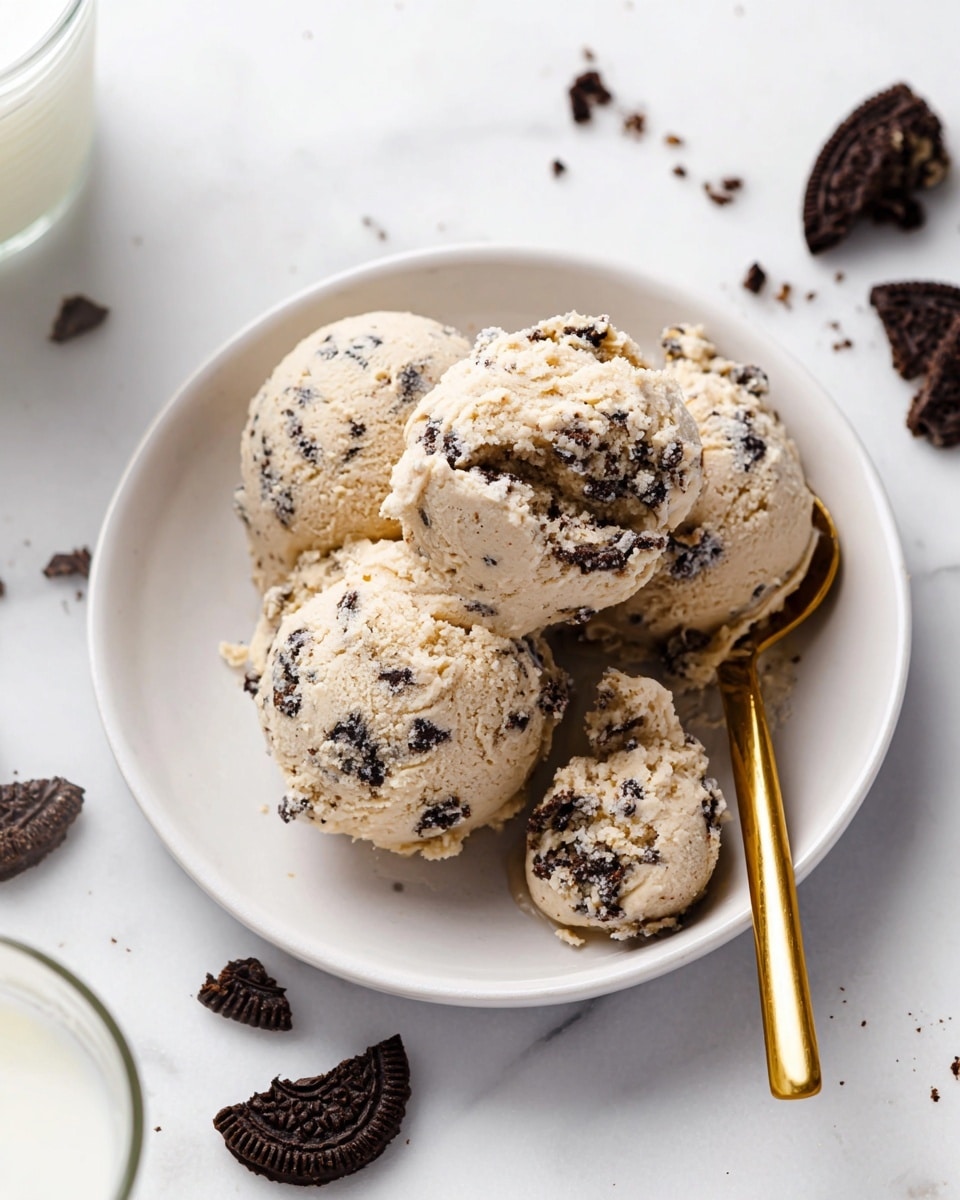 A white plate holds four scoops of light beige cookie dough mixed with dark chocolate cookie bits unevenly spread inside. The dough scoops have a rough, crumbly texture and vary slightly in size, stacked closely together. A gold spoon rests on the right side of the plate with a smaller scoop of dough on it. Around the plate on a white marbled surface are broken pieces of chocolate cookies and a blurred glass of milk is visible at the bottom left. Photo taken with an iphone --ar 4:5 --v 7
