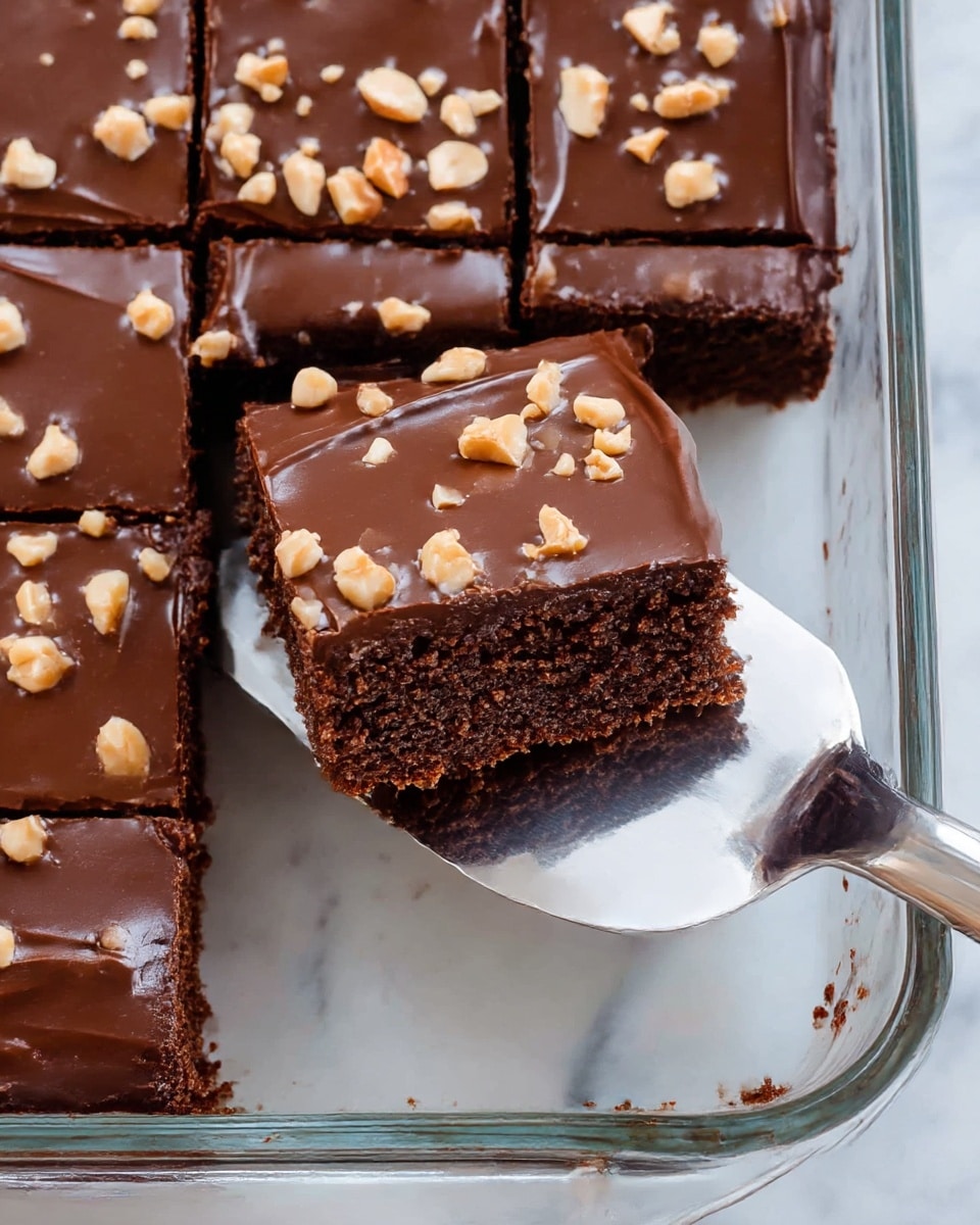 A close-up view shows a chocolate dessert cut into square pieces in a clear glass dish on a white marbled surface. The dessert has two layers: the bottom layer is a moist, dark brown chocolate cake with a soft crumb texture, and the top layer is a smooth, glossy chocolate frosting covered with scattered small light brown chopped nuts. One piece is being lifted by a shiny silver spatula, revealing the thickness and texture of the cake and frosting. photo taken with an iphone --ar 4:5 --v 7