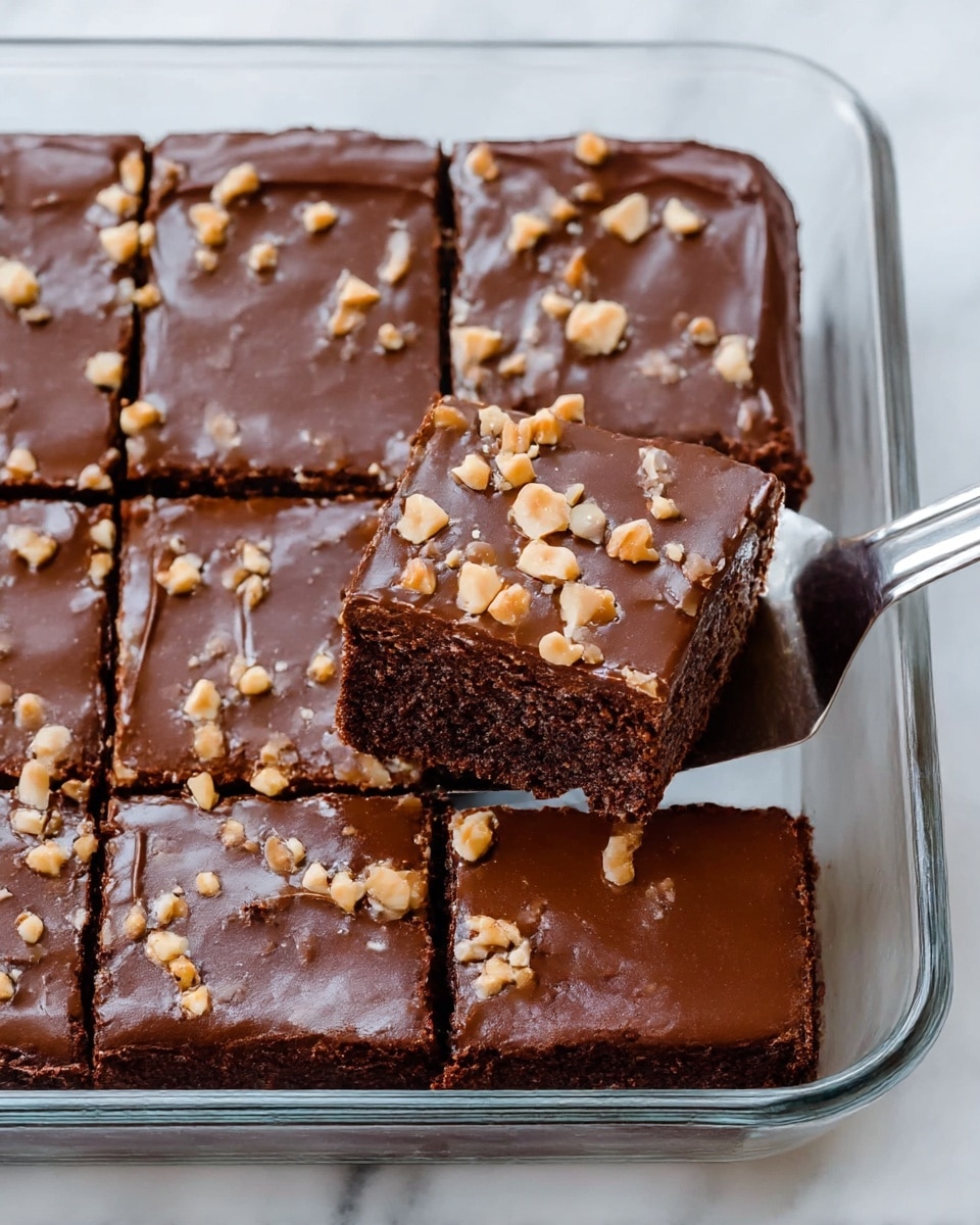 A square chocolate cake cut into smaller squares sits inside a clear glass baking pan on a white marbled surface. The cake has one thick, dark brown layer that looks soft and moist. On top, there is a smooth, glossy chocolate frosting layer spreading evenly across the surface. Light brown chopped peanuts are scattered on top of the frosting. One piece of the cake is being lifted with a shiny metal spatula, showing the cake’s soft texture clearly. Photo taken with an iphone --ar 4:5 --v 7