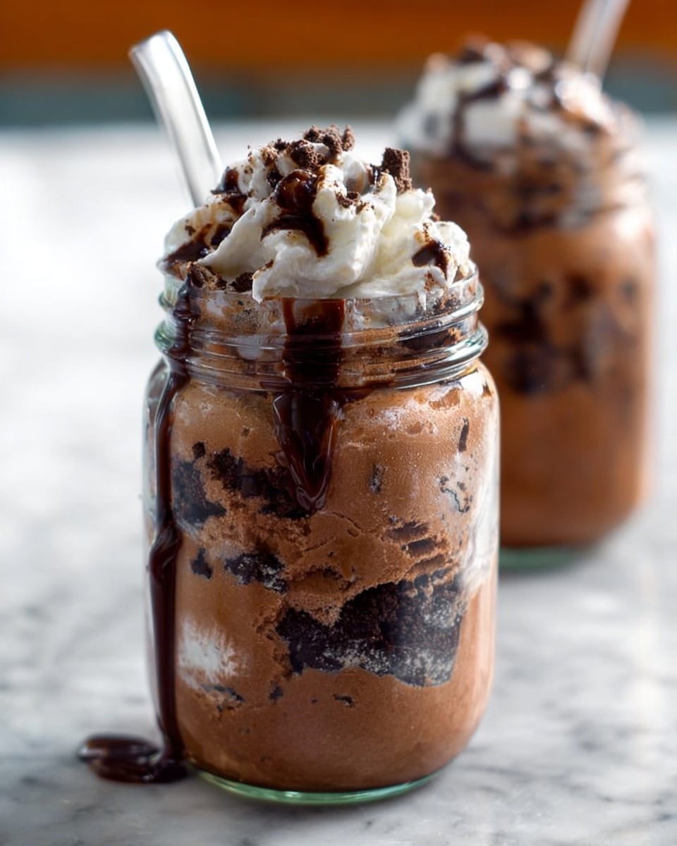 A clear glass jar filled with thick chocolate ice cream mixed with dark chocolate cookie pieces layered unevenly inside. The ice cream has a smooth, creamy texture with bits of crumbly cookies creating dark patches throughout. A generous swirl of white whipped cream tops the jar, drizzled with dark chocolate syrup that drips down the sides of the jar. A white spoon is stuck into the whipped cream, and the jar sits on a white marbled texture. In the background, another similar jar is slightly out of focus, also filled with the same dessert. Photo taken with an iphone --ar 4:5 --v 7