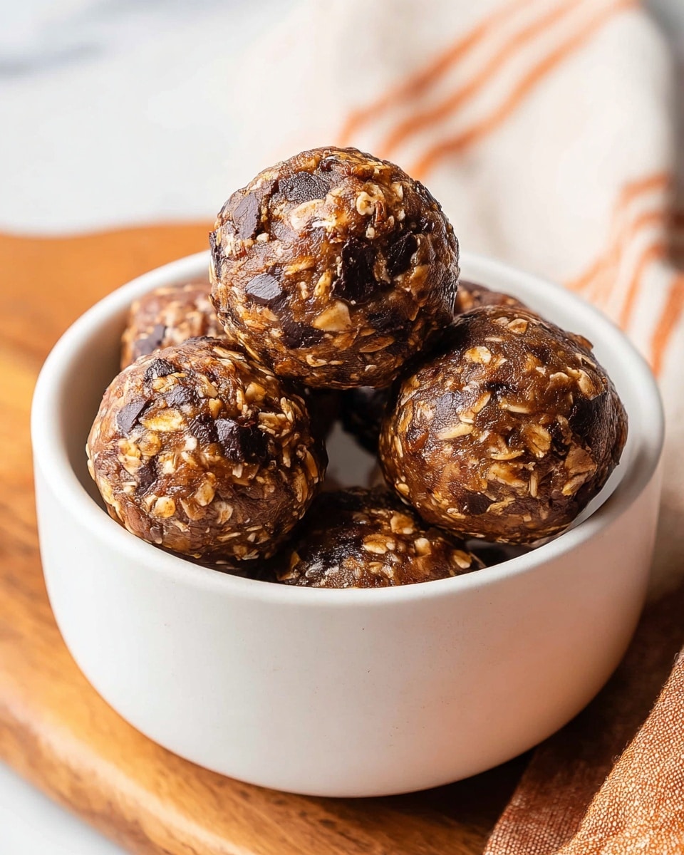 The image shows a white bowl filled with five round energy balls. Each ball has a textured surface with visible pieces of oats and dark chocolate chunks mixed in, giving a marbled look with shades of brown and dark brown. The energy balls appear moist and glossy, stacked neatly inside the bowl. The bowl is placed on a wooden board with an orange-striped cloth next to it, all set against a white marbled surface. photo taken with an iphone --ar 4:5 --v 7