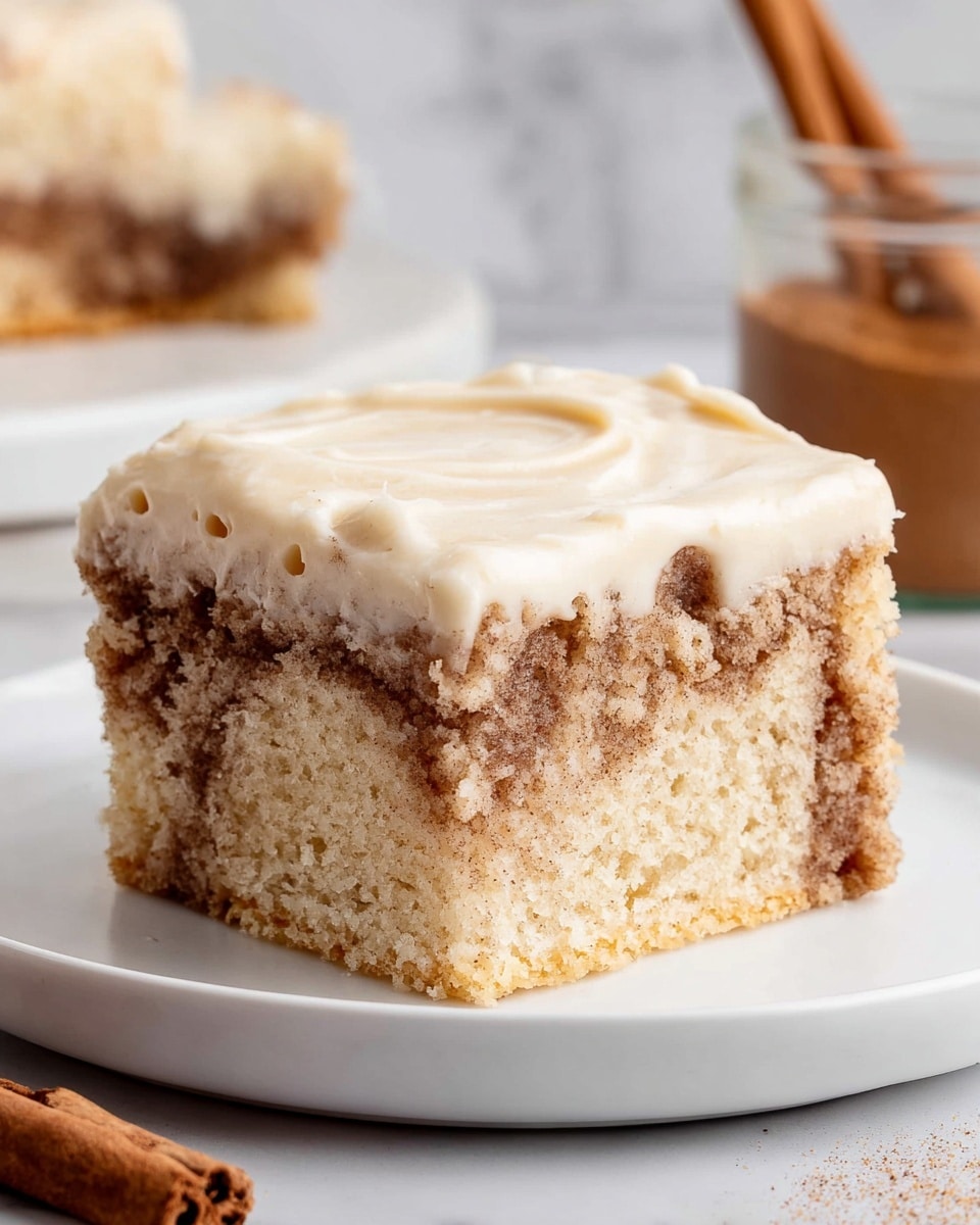 A square piece of cake sits on a white plate, showing two main layers: the bottom layer is a light beige sponge cake with swirls of darker brown cinnamon filling creating a marbled texture, and the top layer is a thick, smooth, creamy off-white frosting gently spread over the cake, slightly dripping down the sides. The background has a white marbled texture with a cinnamon stick and a jar of brown spice blurred seen on the side. Photo taken with an iphone --ar 4:5 --v 7