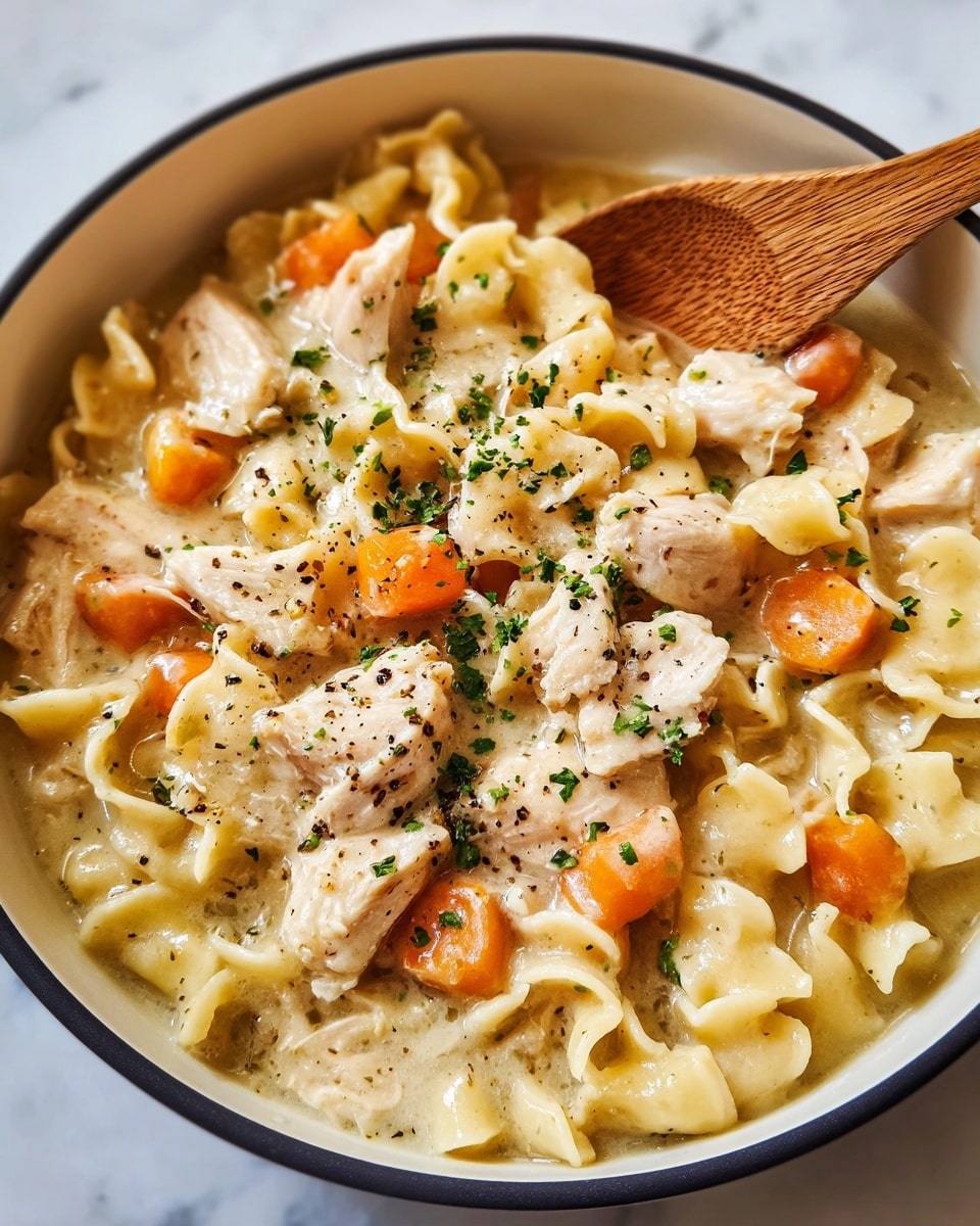 A close-up of a white bowl filled with creamy chicken noodle soup that has three layers: the base is light beige broth with soft pasta pieces shaped like small tubes and ruffled edges, the middle layer has tender chunks of light pink chicken and bright orange carrot cubes, and the top is sprinkled with finely chopped green herbs and specks of black pepper, with a wooden spoon resting on the right side inside the bowl, all set on a white marbled surface. photo taken with an iphone --ar 4:5 --v 7