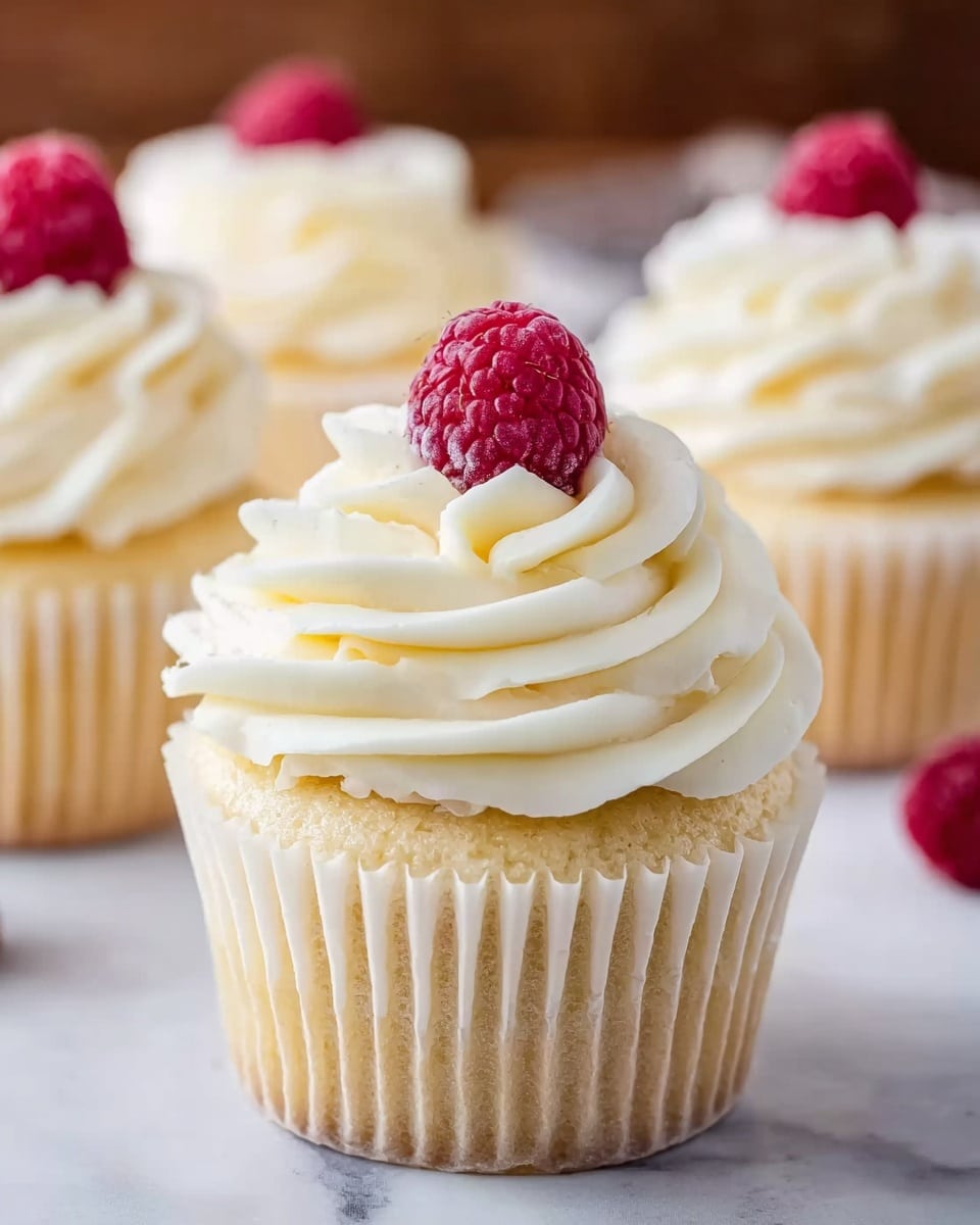 The image shows a close-up of a vanilla cupcake with soft, light yellow cake at the base wrapped in a white paper liner. On top, there is a thick swirl of creamy white frosting, layered in three spirals that create a smooth, fluffy texture. Sitting right in the middle of the frosting is a single bright red raspberry, its bumpy surface standing out against the white frosting. In the background, there are three more similar cupcakes blurred slightly, all on a white marbled surface. photo taken with an iphone --ar 4:5 --v 7