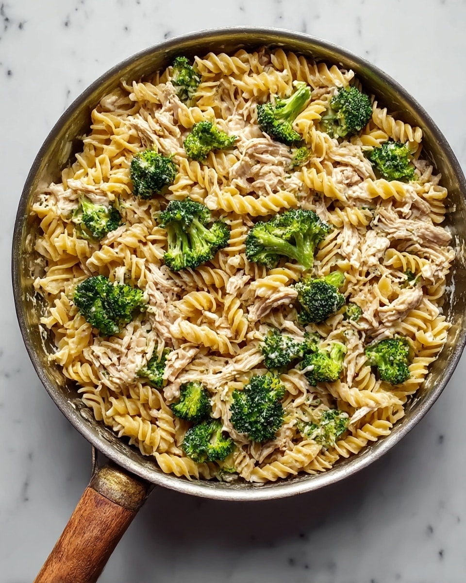 A pan filled with a dish of three layers: the first layer is yellow spiral pasta, the second layer is green broccoli florets placed evenly among the pasta, and the third layer is shredded light brown chicken spread throughout. The textures show the pasta as soft and slightly glossy, the broccoli looks fresh and tender, and the chicken appears moist and fibrous. The pan has a metal body with a wooden handle, and the dish sits on a white marbled surface. photo taken with an iphone --ar 4:5 --v 7