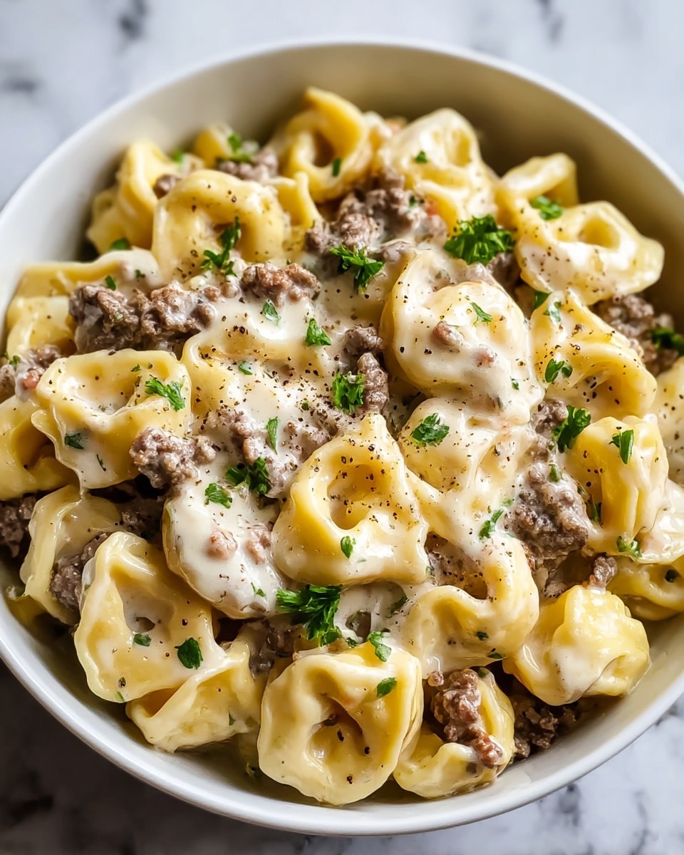 A close-up view of a bowl filled with tortellini pasta that has a light yellow color and a soft texture, mixed evenly with small pieces of browned ground beef. The tortellini and beef are coated in a creamy white sauce with a smooth but slightly thick consistency. Small green herb pieces, likely parsley, are sprinkled on top, adding contrast and freshness. There are also visible specks of ground black pepper scattered throughout. The bowl is white and placed on a white marbled surface. photo taken with an iphone --ar 4:5 --v 7