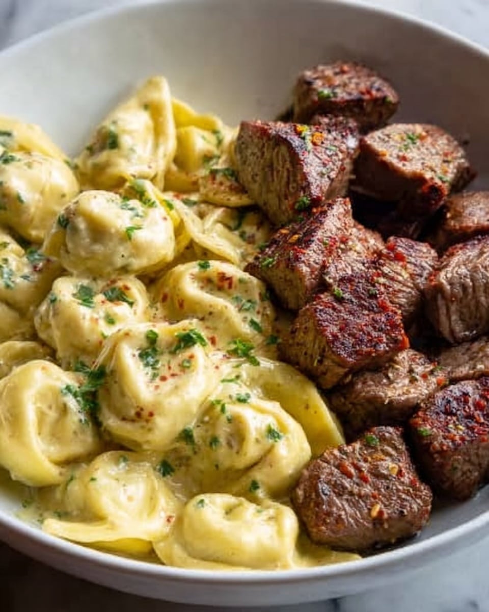 A close-up view of a white bowl showing two main layers: on the left side, a creamy tortellini pasta coated in a smooth pale yellow sauce with small green herb sprinkles on top; on the right side, several pieces of steak cut into cubes with a brown, slightly seared exterior and visible seasoning. The background surface is a white marbled texture. Photo taken with an iphone --ar 4:5 --v 7
