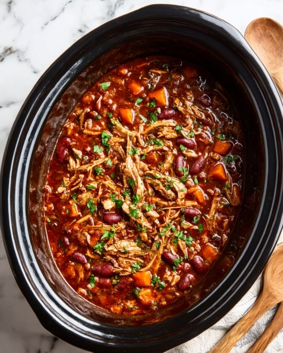 This image shows a black slow cooker filled with a rich, dark red stew that has shredded chicken on top. The stew has visible chunks of vegetables like orange carrots and red beans mixed in with the thick sauce. The shredded chicken is spread evenly across the surface and is garnished with green herbs. The cooker is placed on a white marbled surface with part of a striped linen cloth and wooden spoons visible nearby. Photo taken with an iphone --ar 4:5 --v 7