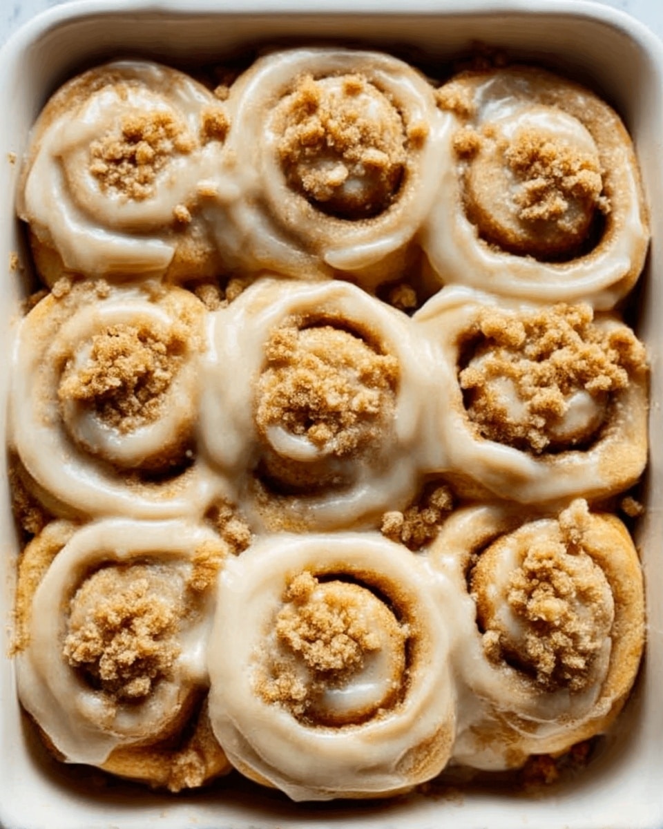 The image shows a white dish filled with nine cinnamon rolls arranged in three rows of three. Each roll has a light golden-brown color on the outside with visible swirls of dough spiraled inward. The rolls are covered in a creamy light tan glaze that makes them look soft and sticky. On top of several rolls, there are small clusters of crumbly, light brown streusel. The dish is placed on a white marbled texture surface. Photo taken with an iphone --ar 4:5 --v 7
